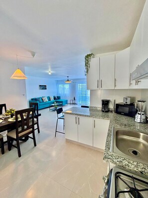 A modern kitchen with white cabinets, a granite countertop, and stainless steel appliances. There is a dining area with a table and chairs, and a blue sofa in the background. - 2-bedroom  spacious apartment in tranquil Punta Cana, very close to the mall (Punta Cana)
