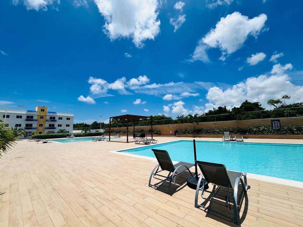 A swimming pool with lounge chairs, a covered seating area, and a building in the background.
