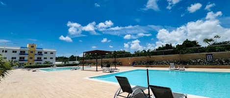 A swimming pool with lounge chairs, a covered seating area, and a building in the background.