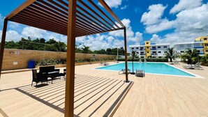 Ein Poolbereich mit einer Holzpergola, Liegestühlen und Blick auf ein Hotel im Hintergrund.