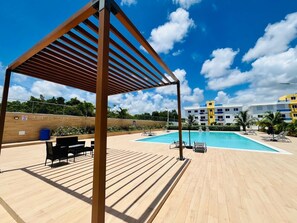 A pool area with a wooden pergola, lounge chairs, and a view of a hotel building in the background. - 2-bedroom  spacious apartment in tranquil Punta Cana, very close to the mall (Punta Cana)