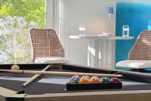 A pool table with balls, a cue stick, and two wicker chairs.