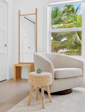 A modern living room with a sofa, a small stool, a wooden bench, and a large window with a view of palm trees.