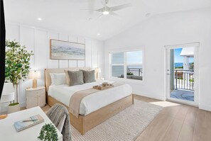 Ein Schlafzimmer mit Bett, Nachttischen, einem Schreibtisch, einem Stuhl, einer Pflanze, einem Fenster und einem Balkon mit Aussicht.