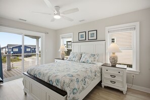 A bedroom with a bed, two bedside tables, a ceiling fan, and a view of the outside. - Pelican Parade: Ocean View, Pet-Friendly 5 Bedroom Luxury Retreat with Private Pool and Elevator (Oak Island)