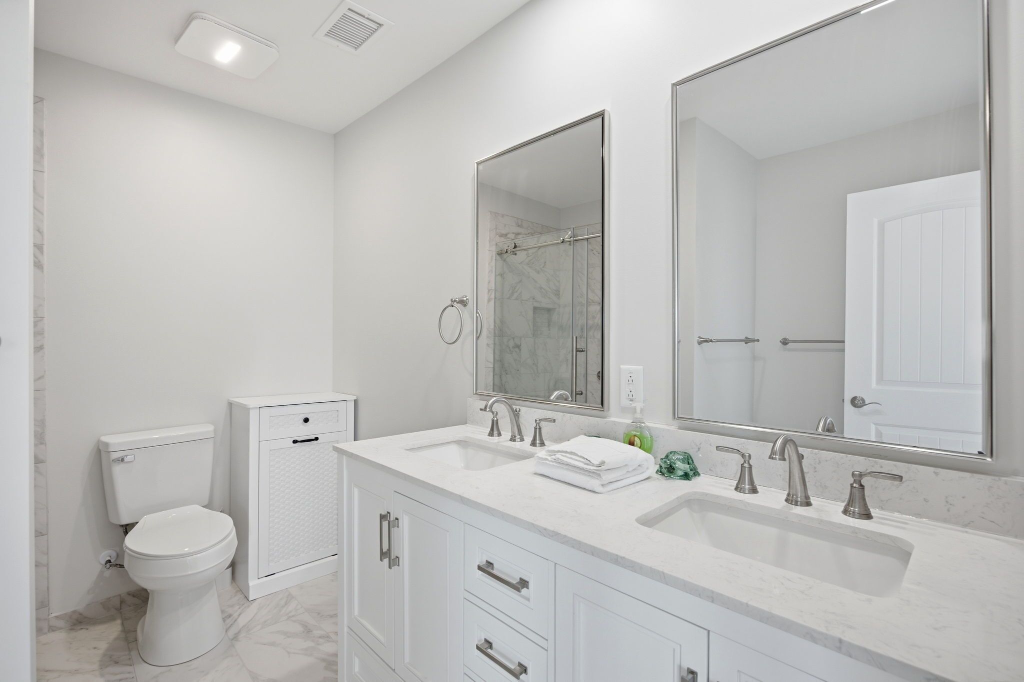 A modern bathroom with a white toilet, a shower enclosure, a double sink vanity with cabinets, and a large mirror.