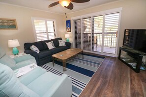 Ein Wohnzimmer mit einer Couch, einem Couchtisch und einem Fernseher, einem Deckenventilator und Blick durch eine Schiebetür.