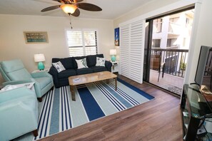 A living room with a sofa, armchairs, a coffee table, and a television. There is a ceiling fan, a window with blinds, and a sliding glass door leading to an outdoor area. - Newly Remodeled Condo Ocean Village Club F26 (St. Augustine)