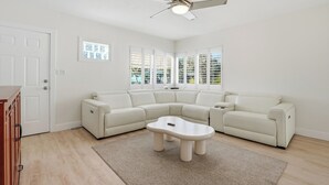 Un salon moderne avec un canapé d’angle, une table basse, un ventilateur de plafond et de grandes fenêtres.