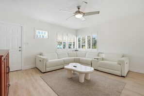Ein modernes Wohnzimmer mit einem Ecksofa, einem Couchtisch, einem Deckenventilator und großen Fenstern.