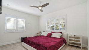 Une chambre à coucher avec un lit, des tables de chevet blanches, un ventilateur de plafond et des fenêtres équipées de stores.