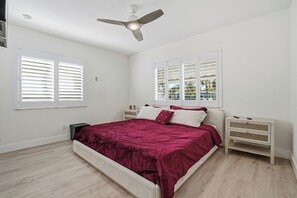 Ein Schlafzimmer mit einem Bett, weißen Nachttischen, einem Deckenventilator und Fenstern mit Jalousien.