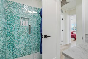 Un baño con cabina de ducha de vidrio, una puerta blanca y una cama roja en la habitación contigua.