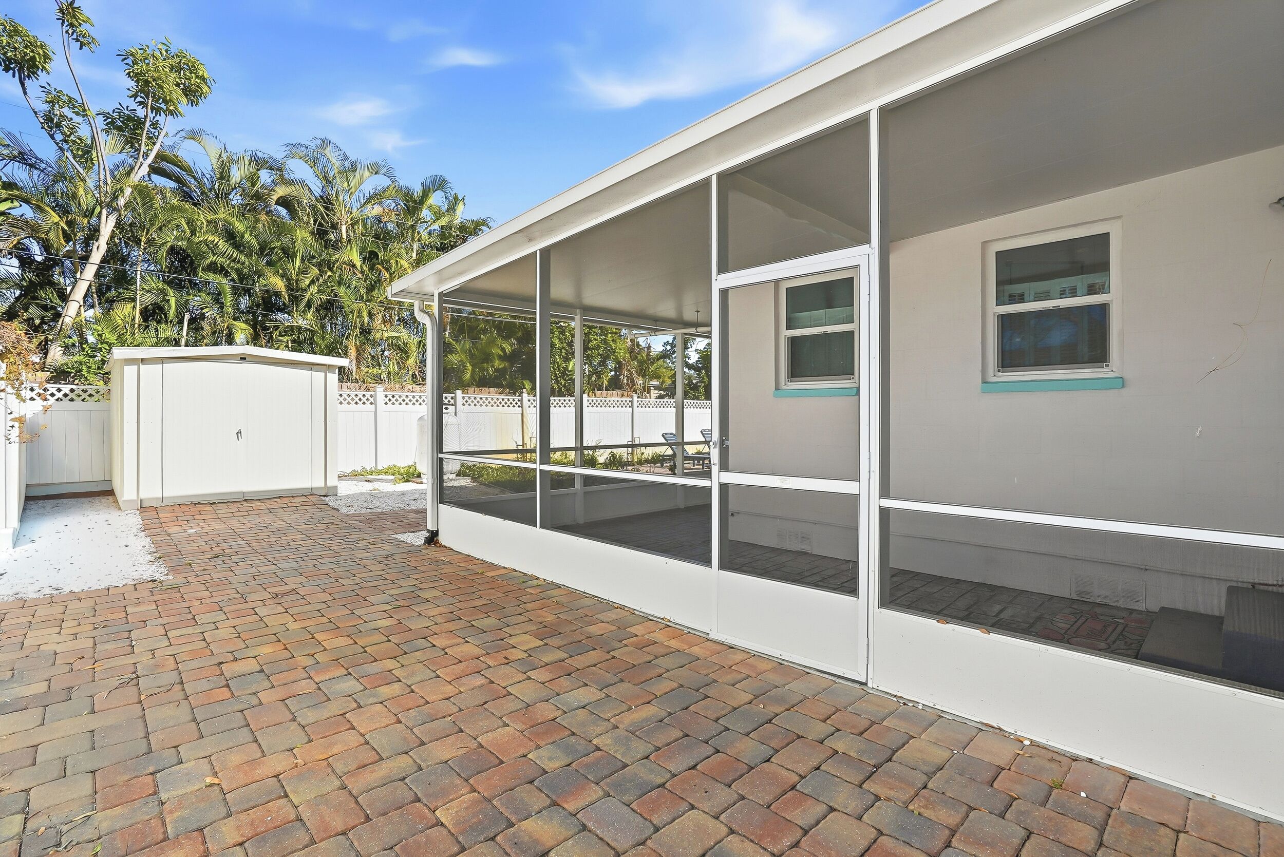 A covered outdoor area with a brick floor, a white building with multiple windows, and a white fence.
