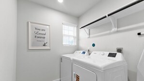 A laundry room with two white washers and dryers, a framed inspirational quote, and a window with blinds. - Brand New 3BR Home with Game Room & BBQ Near The Woodlands (Decker Prarie)