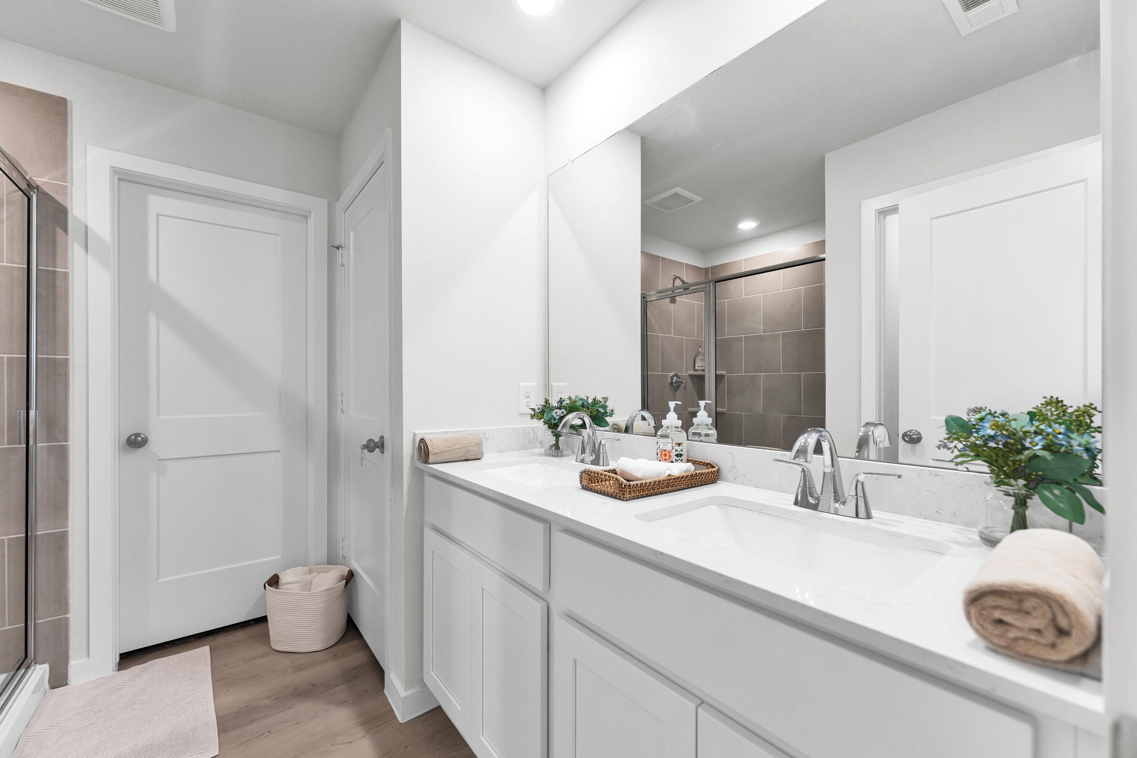 A modern bathroom with a large mirror, two sinks, a shower, and a walk-in closet.