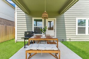 Eine Terrasse mit Holztisch und Bank, einer hängenden Lampe und einem Grill vor einem Haus.