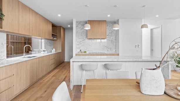 A modern kitchen with wooden cabinets, a marble countertop, pendant lighting, and a dining area with a white table and chairs.