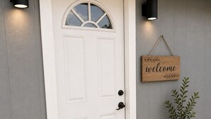 Uma porta branca com um janelão semicircular, uma placa de boas-vindas, um vaso de planta e um tapete de entrada.