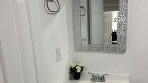 Um banheiro com pia branca, espelho com moldura de mosaico, porta-papel higiênico e porta.