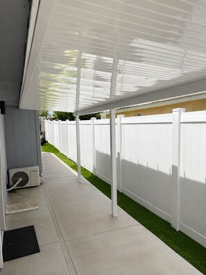 Ein überdachter Patio-Bereich mit weißem Zaun, einem Betonweg und einem kleinen Rasenstück.