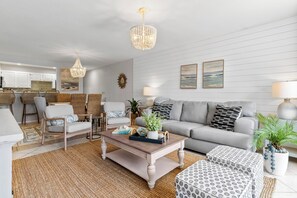 A living room with a sofa, coffee table, and chairs, a kitchen in the background, and a chandelier. - La Mirage 102: Spacious 3-bedroom condo in Port Aransas, perfect for family getaways! (Port Aransas)