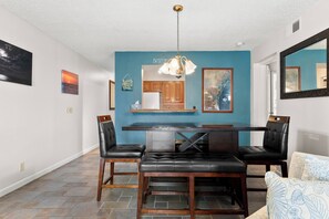 A dining area with a blue accent wall, a kitchen in the background, and a chandelier. - Discover La Mirage 224: a cozy 2-bedroom condo with partial Gulf views in Port Aransas. (Port Aransas)