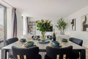 Un coin repas avec une table dressée pour quatre personnes, entourée de chaises, un vase rempli de fleurs et une bouteille de champagne.