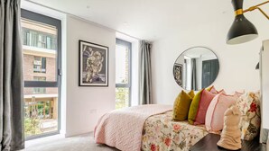 Ein Schlafzimmer mit großem Fenster, einem Bett mit floralem Bettbezug, einem runden Spiegel und einer schwarzen Schreibtischlampe.