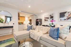 A modern living room with a sofa, coffee table, and a kitchen area in the background. - The London Dock HQ - 3 Bed with AC for 6 Guests (London)