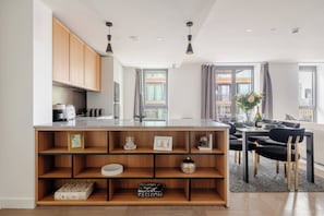 A modern kitchen with a central island, wooden shelving, and a dining area with black chairs and a table. - The London Dock HQ - 3 Bed with AC for 6 Guests (London)