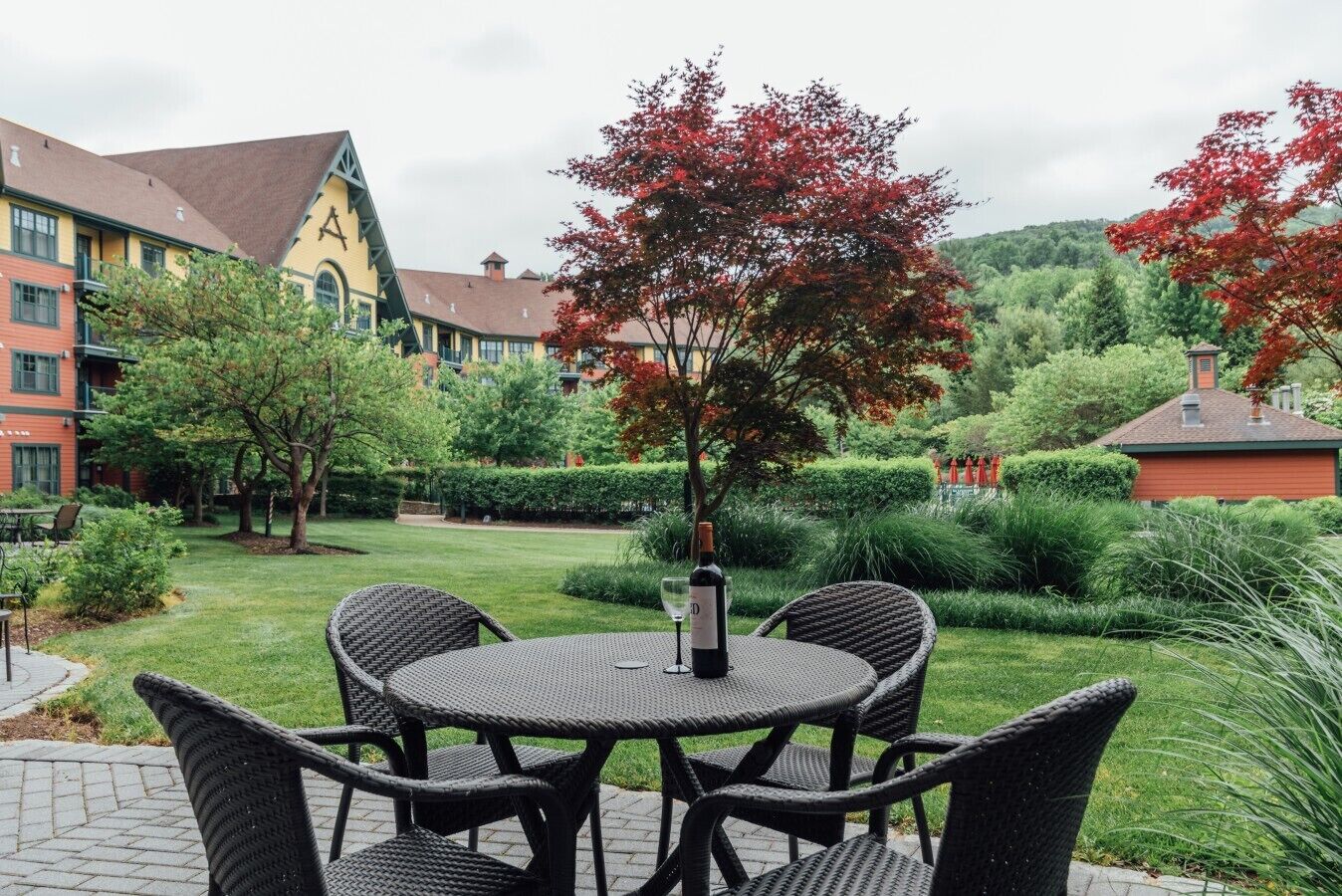 An outdoor seating area with wicker chairs and a table, surrounded by well-maintained greenery and a building with a distinctive roof.