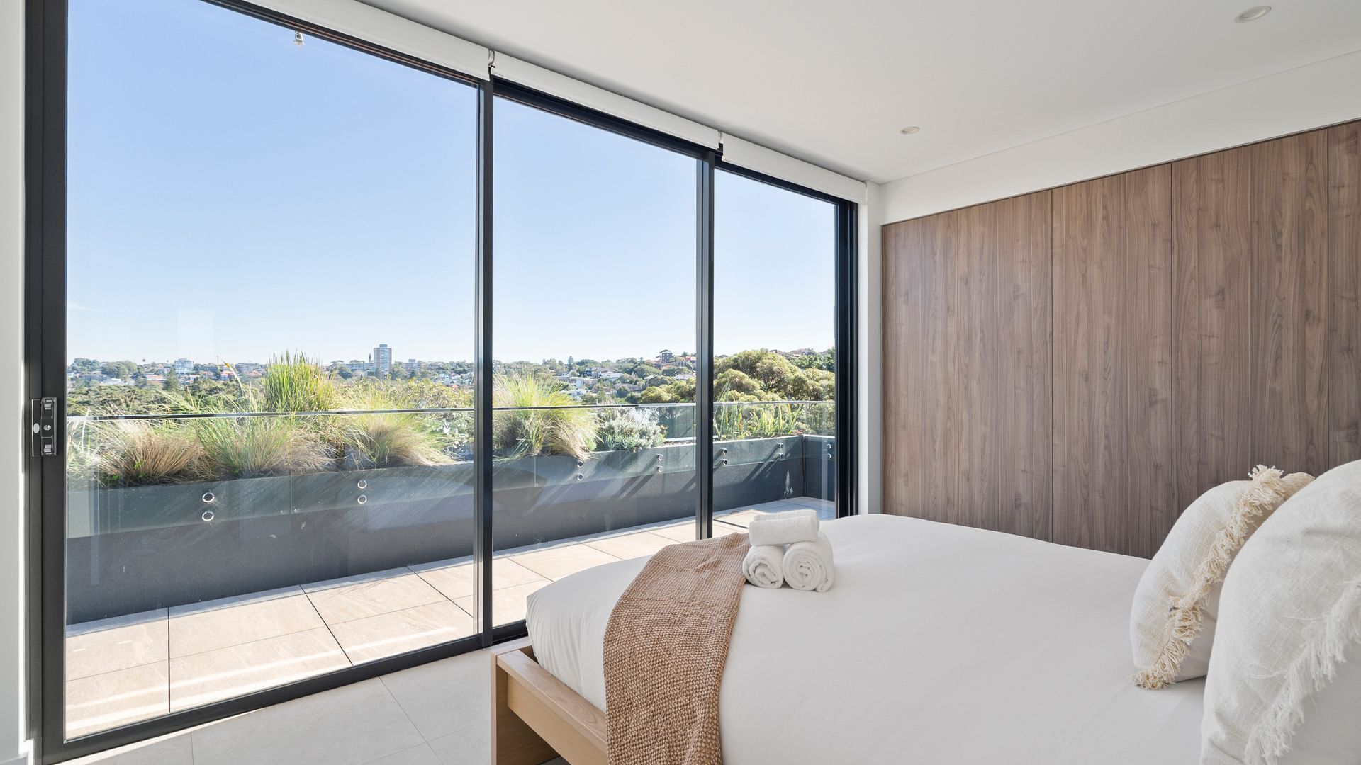 A bedroom with a large sliding glass door, a bed with white linens, a wooden headboard, and a view of a cityscape.