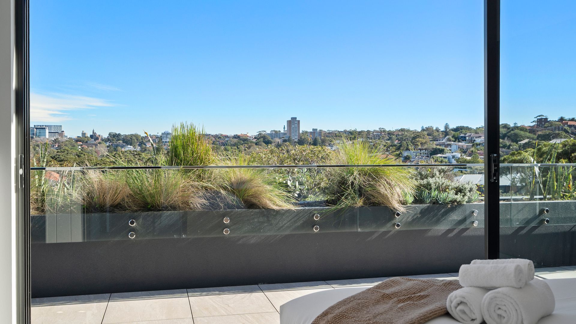 A balcony with a view of a cityscape, a glass railing, and a small seating area with towels.