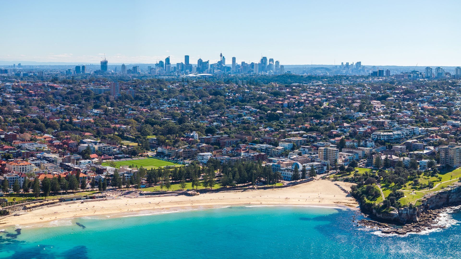 Une vue aérienne d’une ville côtière, avec une plage, des quartiers résidentiels et une silhouette urbaine formée par des bâtiments.