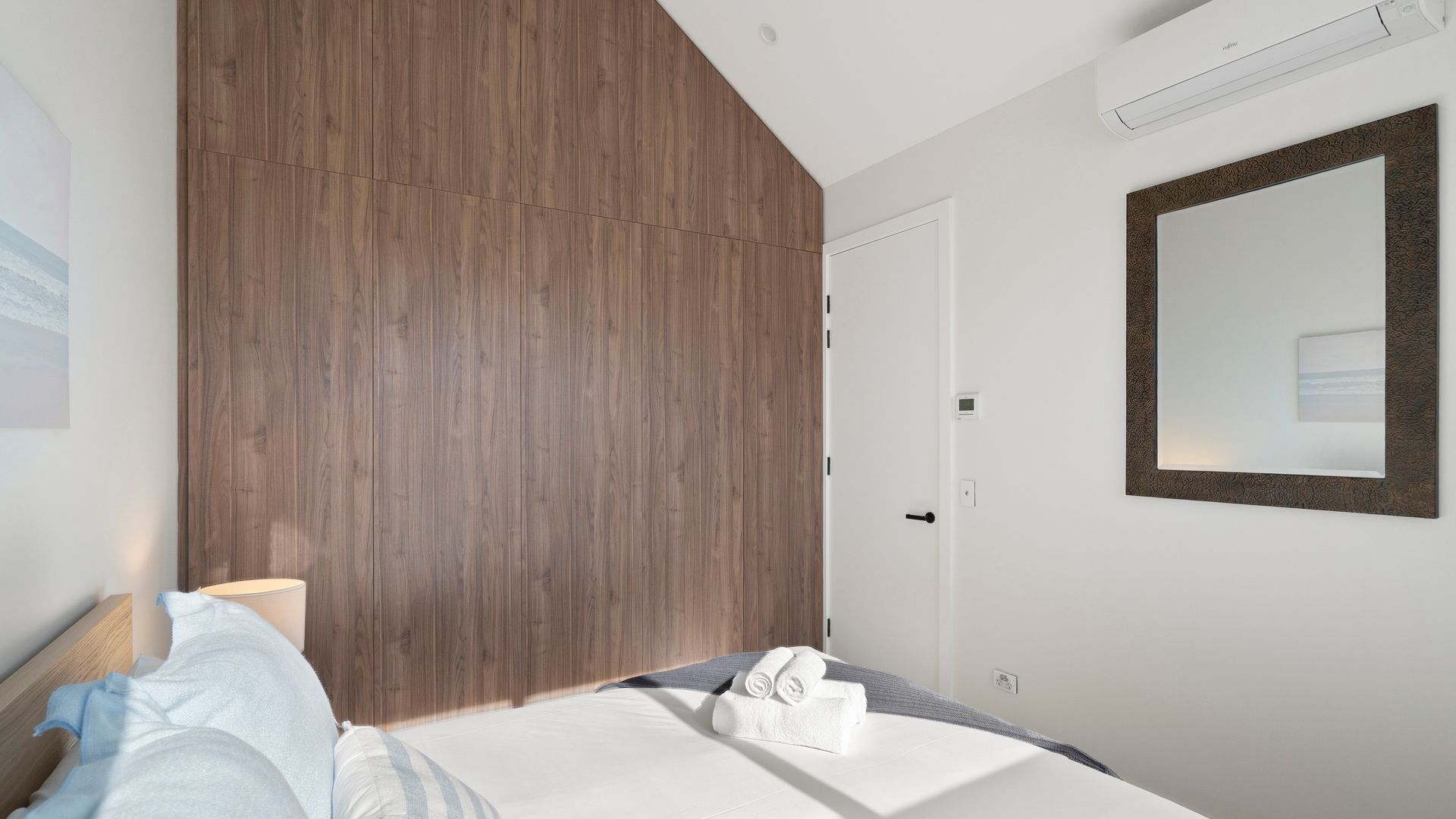 A modern hotel room with a wooden headboard, a bed with white linens, a large mirror, and a thermostat on the wall.