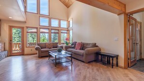 A spacious living room with a high ceiling, large windows, and a wooden floor. - New- Cozy Pathway Pines (Truckee)