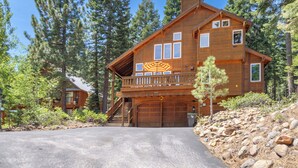 A two-story wooden cabin with a balcony, a garage, and a driveway leading to the entrance. - New- Cozy Pathway Pines (Truckee)