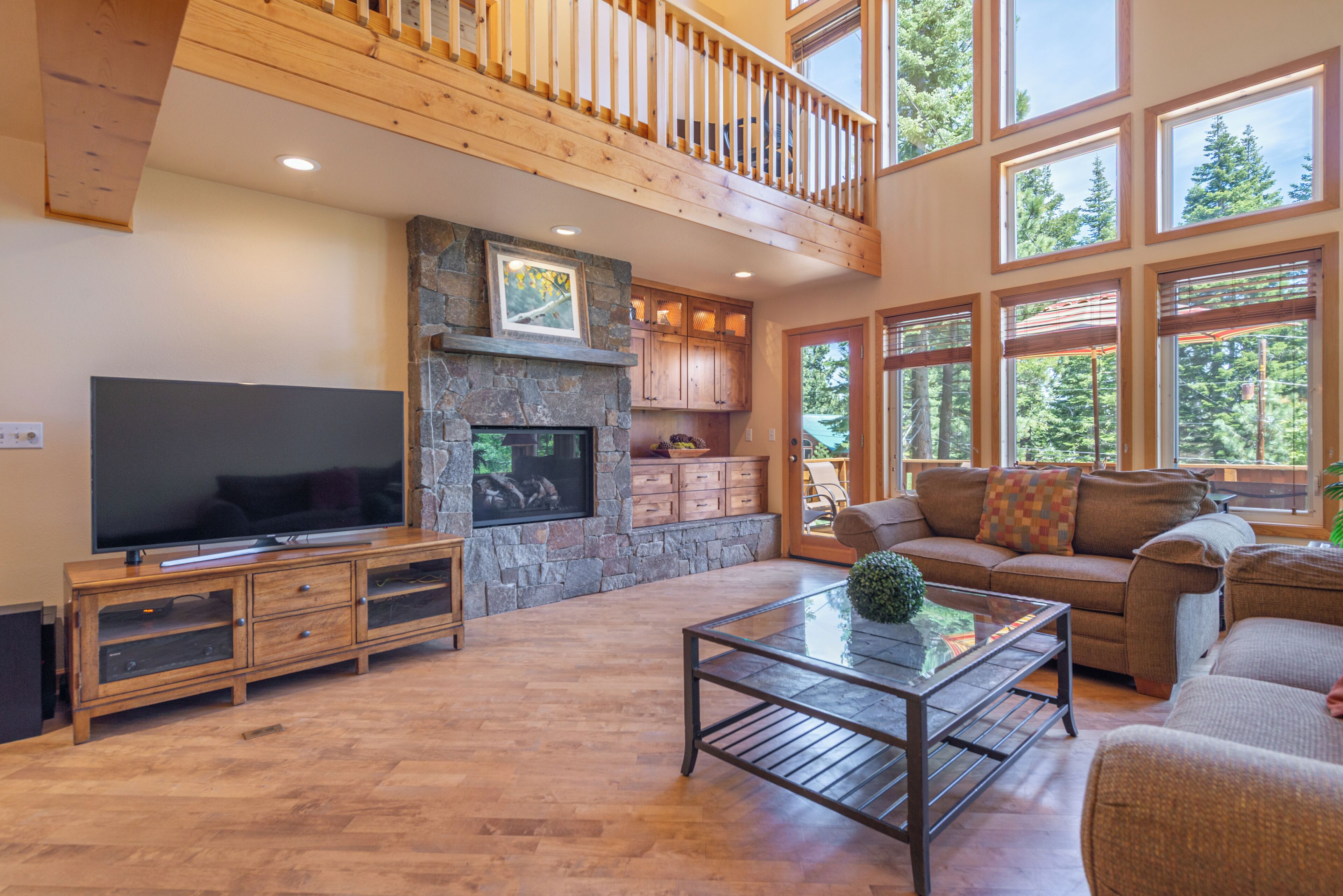A spacious living room with a stone fireplace, a television, and a large window overlooking a forest.