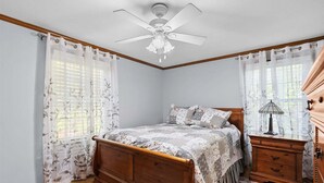 Un dormitorio con cama de madera, una mesita de noche, lámpara, ventilador de techo y cortinas traslúcidas.