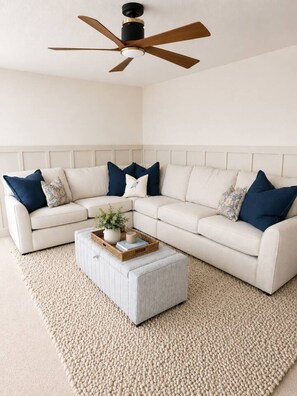 Una sala de estar con un sofá sectional blanco, un pequeño otoman, un ventilador de techo y una alfombra con textura.