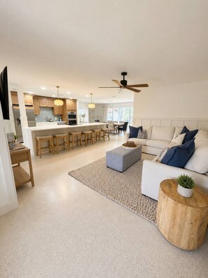 A modern kitchen with a central island, a dining area with chairs, a living room with a sofa and ottoman, and a ceiling fan.