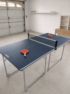 A ping pong table with two paddles, a garage door, and a wooden cabinet in a room.
