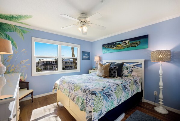 A bedroom with a bed, a window, a ceiling fan, a nightstand, a lamp, and a painting on the wall. - Direct Beachfront | Coastal 3BR Retreat (Bradenton Beach)
