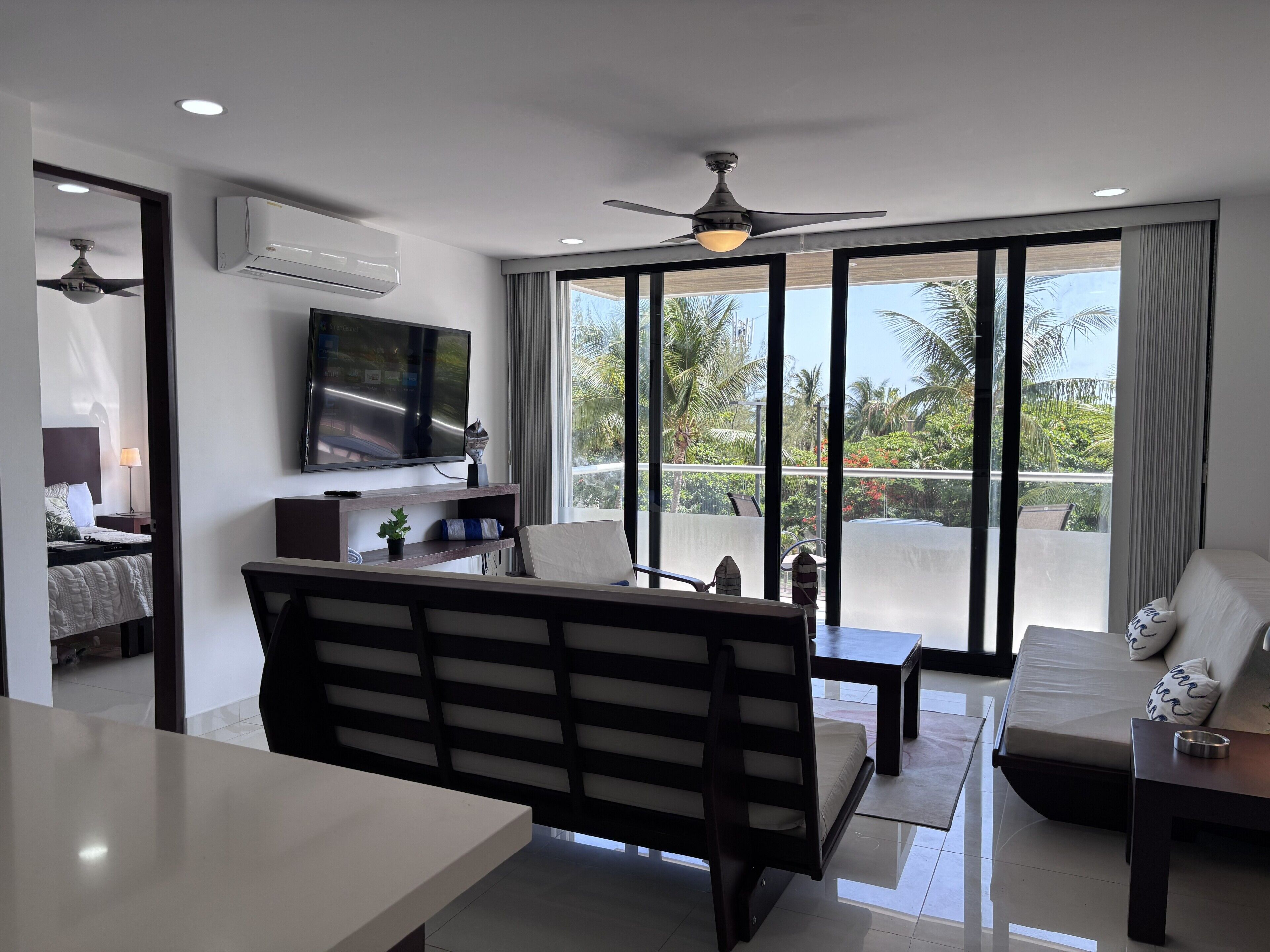 Een moderne woonkamer met een flatscreen-tv, een plafondventilator en een balkon met uitzicht op palmbomen.