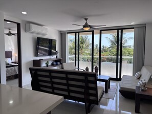 A modern living room with a flat-screen TV, a ceiling fan, and a balcony with a view of palm trees. - Delightful 2 bedroom Condo & Ocean View Rooftop& just across the beach (Cancún)
