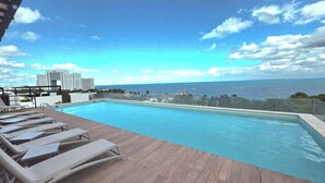 Piscina com espreguiçadeiras e vista para o oceano e um edifício ao longe.