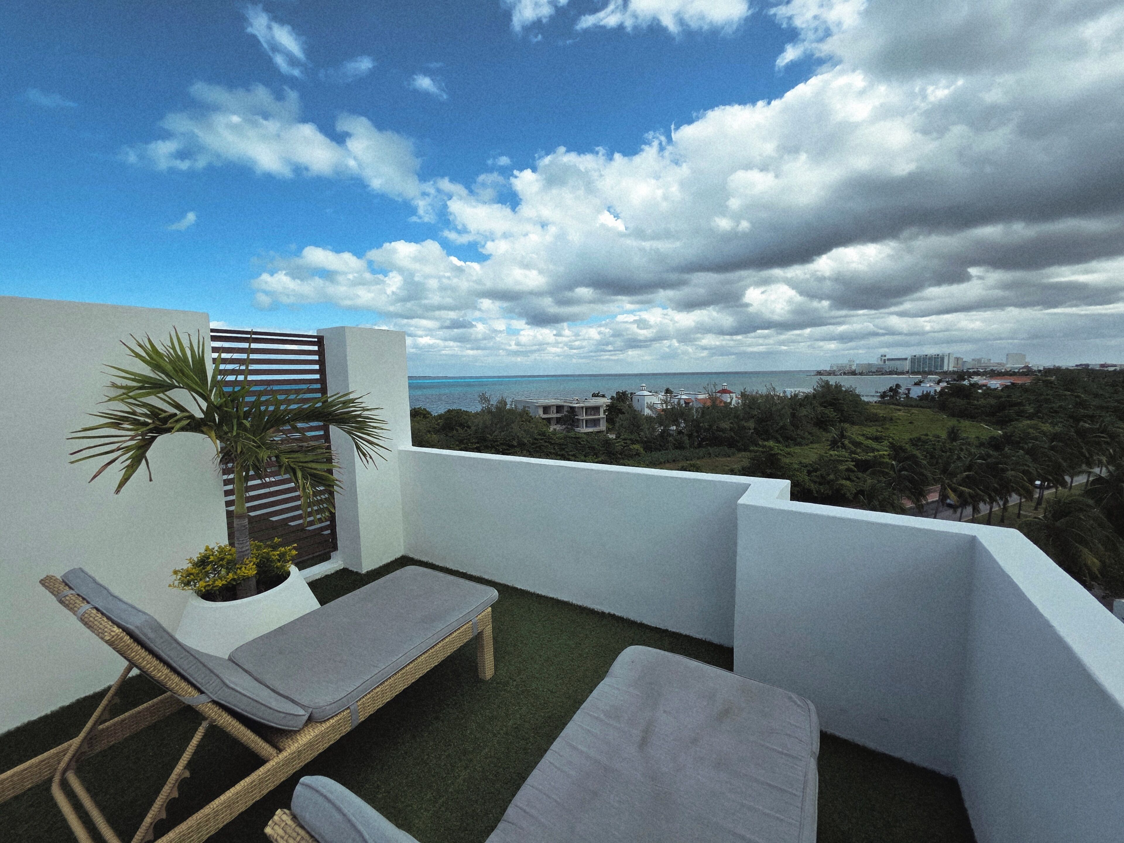 A balcony with two lounge chairs, a small palm tree, and a view of the sea and cityscape.