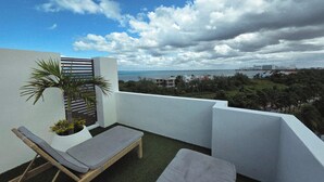 Varanda com duas poltronas de descanso, uma pequena palmeira e vista para o mar e a paisagem urbana.