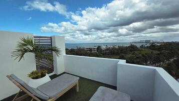 A balcony with two lounge chairs, a small palm tree, and a view of the sea and cityscape.